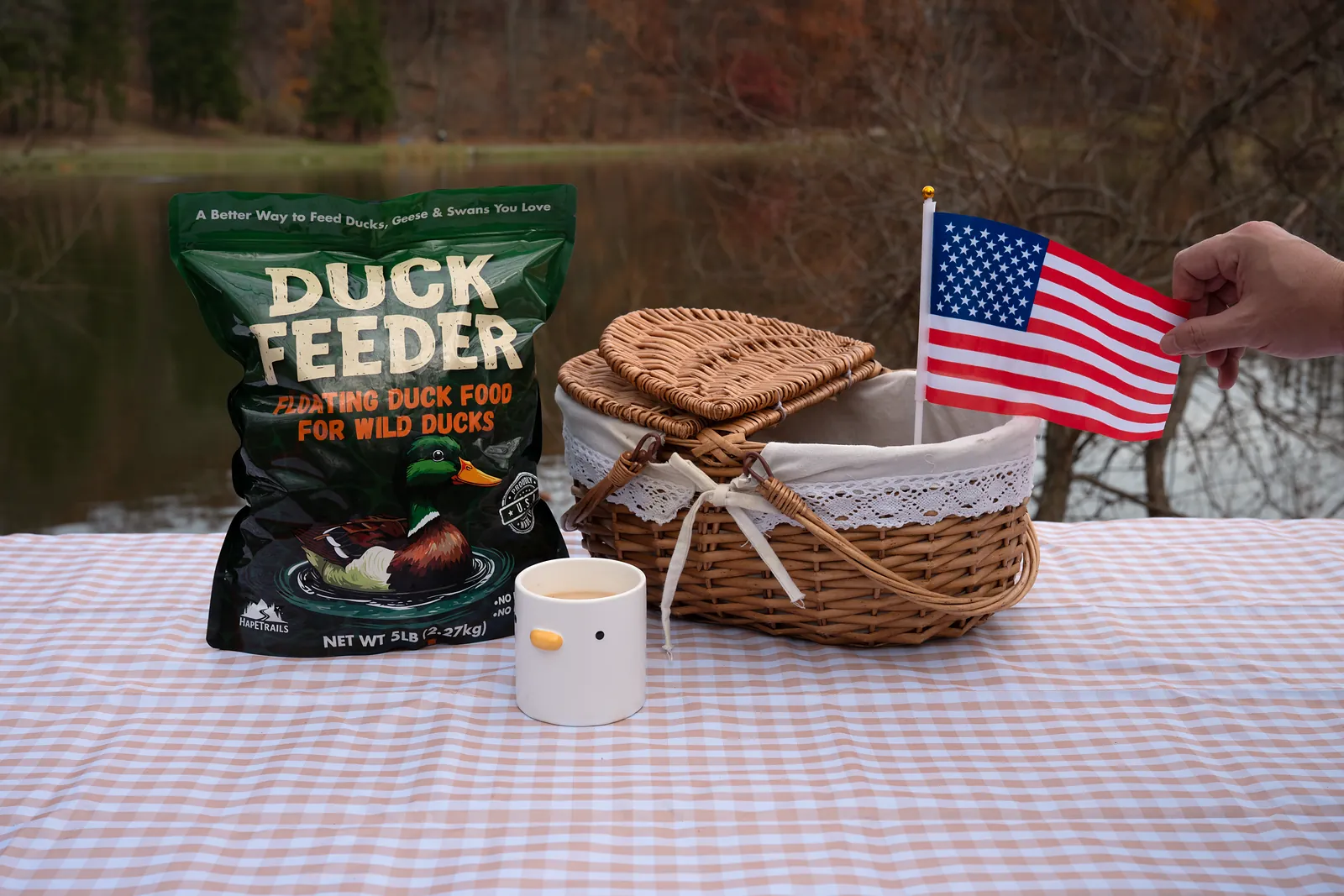 A Duck Feeder bag next to a picnic basket and a small American flag on a checkered tablecloth by the pond
